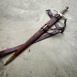 May include: A sword with a metallic crossguard and pommel, housed in a brown leather scabbard featuring intricate embossed knotwork patterns. The scabbard is tipped with a brass chape and is attached to a dark brown leather belt with metal buckles.