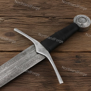 Può includere: Una spada con lama in acciaio Damasco a motivi, guardia in metallo color argento, impugnatura rivestita in pelle nera e pomolo circolare color argento con centro testurizzato. Il testo NorthlandForge è visibile nell'immagine.