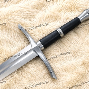 Puede incluir: Una espada de metal plateado con una hoja recta de doble filo, una guarda con gavilanes ligeramente curvados y una empuñadura envuelta en negro. El pomo es redondeado y metálico, a juego con la guarda y la hoja.