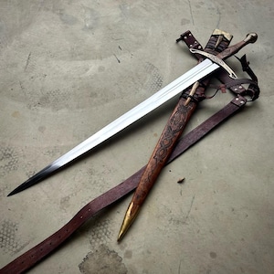 May include: A steel sword with a cross-shaped guard and a metallic pommel, accompanied by a brown leather scabbard featuring intricate embossed knotwork patterns, a brass tip, and attached leather straps for carrying the weapon securely.