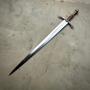 May include: A long, double-edged steel sword featuring a pointed tip, a central fuller, and a crossguard with decorative engravings. The handle is wrapped in brown material and terminates in a rounded metal pommel at the base of the grip.