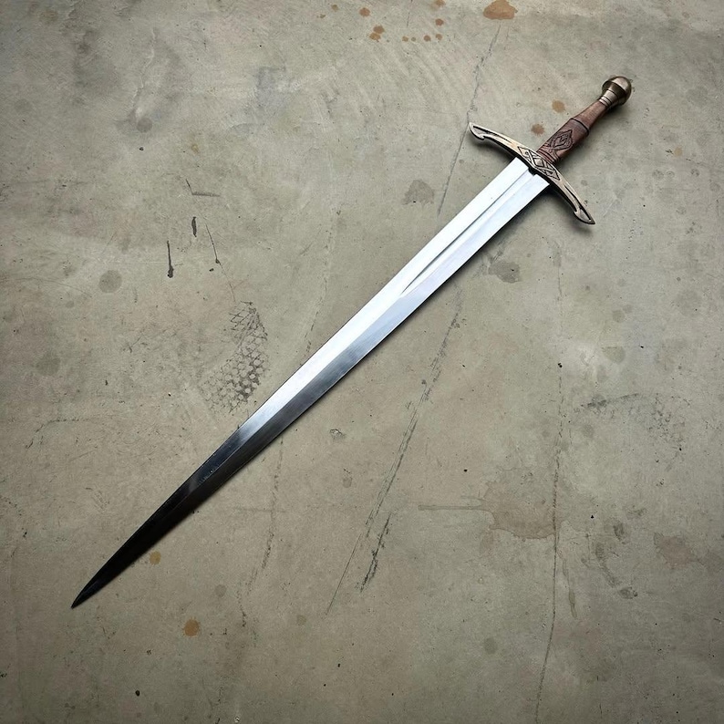 May include: A long, double-edged steel sword featuring a pointed tip, a central fuller, and a crossguard with decorative engravings. The handle is wrapped in brown material and terminates in a rounded metal pommel at the base of the grip.