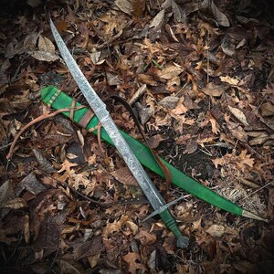 May include: A long sword with a patterned Damascus steel blade, a green leather-wrapped hilt, and a matching green leather scabbard featuring brown leather straps and a metal tip, resting diagonally across a bed of fallen brown autumn leaves.