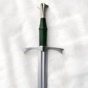 Puede incluir: Una espada de acero con hoja de doble filo, una guarda de metal plateado y una empuñadura envuelta en color verde oscuro. El pomo es una pieza de metal plateado con un diseño angular y ensanchado que combina con el acabado metálico.