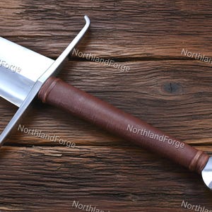 May include: A sword with a polished silver-colored metal blade, a straight crossguard, and a brown leather-wrapped grip. The hilt features a rounded, metallic pommel. The text NorthlandForge is visible multiple times across the image.