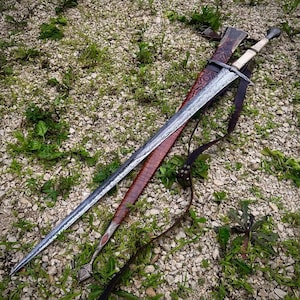 Op de afbeelding: Een lang zwaard met een stalen lemmet met patroon, een lichtgekleurd handvat en een pareerstang. Het zwaard rust op een bruine lederen schede met reliëfpatronen, een metalen kap aan de punt en bevestigde donkere lederen draagriemen.