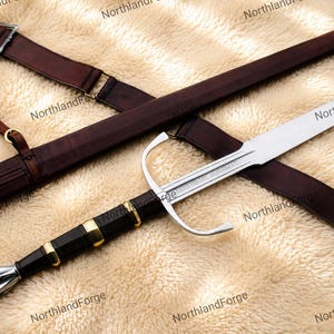 May include: A steel sword with a silver-colored pommel, a black grip with three gold-colored rings, and a silver crossguard. The sword is accompanied by a dark brown leather scabbard featuring adjustable straps and metal buckles. NorthlandForge text.