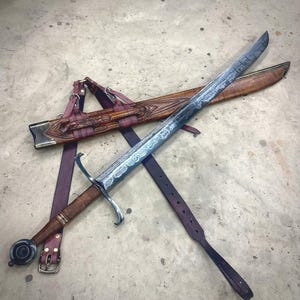 May include: A long sword with a patterned Damascus steel blade, a brown leather-wrapped hilt, and a circular pommel. It is accompanied by a matching brown carved wooden scabbard featuring intricate detailing and a dark purple leather belt system.