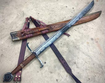 May include: A long sword with a patterned Damascus steel blade, a brown leather-wrapped hilt, and a circular pommel. It is accompanied by a matching brown carved wooden scabbard featuring intricate detailing and a dark purple leather belt system.