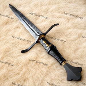 May include: A dagger featuring a patterned Damascus steel blade, a black handle with a gold-colored fleur-de-lis emblem, and a black crossguard. The pommel is black and flared, and the handle includes a gold-colored band near the base.