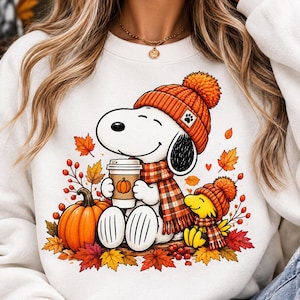 Puede incluir: Una sudadera blanca de cuello redondo con un gráfico de Snoopy y Woodstock usando gorros de punto naranja y bufandas a cuadros. Snoopy sostiene un vaso de café con un icono de calabaza, rodeado de hojas de otoño, bayas y una calabaza naranja.