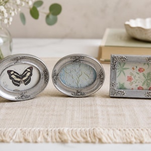 May include: Three small picture frames with a metallic silver finish and ornate, raised floral detailing. Two frames are oval-shaped, one containing a butterfly illustration and the other a floral print. The third frame is rectangular with a floral pattern.