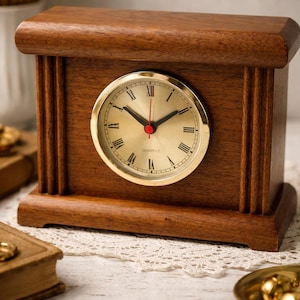 May include: A wooden quartz clock with a gold-colored face and Roman numerals. The clock has a rectangular shape and a dark brown finish. The clock's hands are black, with a red second hand.