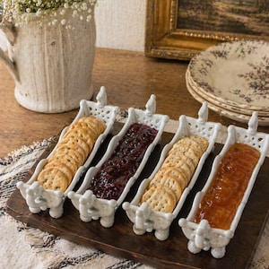 Puede incluir: Una bandeja de madera con cuatro fuentes de cerámica blanca. Dos fuentes contienen galletas saladas, y las otras dos están llenas de mermelada roja oscura y mermelada de naranja. La bandeja está sobre un paño estampado.