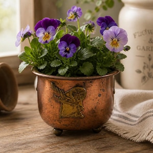 May include: A copper-colored planter pot with a decorative emblem, filled with vibrant purple and yellow pansies. The pot sits on a wooden surface, with a stack of terracotta pots and a ceramic jar in the background. The flowers are in full bloom.