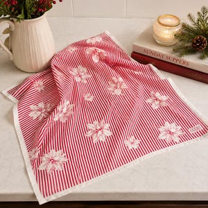 May include: A square kitchen towel featuring a pattern of thin vertical red and white stripes with a repeating white poinsettia floral print. The fabric has a white border and a small white tag in the corner with the text CLUB7 printed in black.