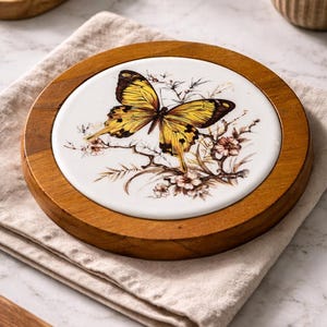 May include: Round wooden coaster with a white ceramic center featuring a detailed illustration of a yellow and brown butterfly. The butterfly is surrounded by floral accents. The coaster is set on a beige linen napkin.