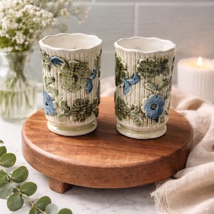 May include: Two ceramic vases with a floral design in green and blue, set on a round wooden tray. The vases have a textured, vertical striped pattern. A candle and flowers are in the background.