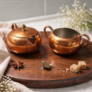 May include: A copper tea set with a small teapot and a sugar bowl, displayed on a wooden tray. A small brass bird figurine, star anise, and sugar cubes are also on the tray. The set is placed on a wooden surface.