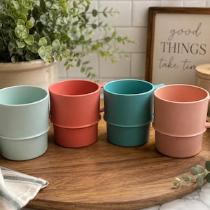 May include: A set of four cylindrical mugs in pastel mint, coral, teal, and light pink. Each mug features a horizontal ridge around the center and a square handle. The mugs are arranged in a row on a round wooden tray, showcasing a smooth matte finish and a stackable design structure.