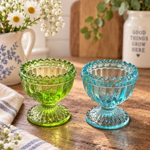 May include: Two glass pedestal bowls featuring a scalloped rim and a beaded edge design. One bowl is vibrant lime green, and the other is a clear light blue. Both bowls have a vertical ribbed pattern on the exterior and a circular base.