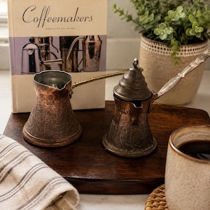 May include: Two copper coffee pots with long handles and intricate embossed floral patterns. One pot is open, while the other features a conical lid. They are positioned in front of a book titled Coffeemakers Baristas at Home.