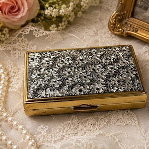 May include: A rectangular metal case featuring a gold-toned frame and a black lid with an intricate, raised silver-colored floral and scrollwork pattern. The compact box has a hinged closure and a smooth, polished finish on the side edges.