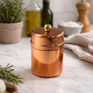 May include: A copper-colored canister with a lid and a small gold-colored knob. The canister is cylindrical and sits on a marble countertop. Other kitchen items are in the background, including a small potted plant and bottles of oil.