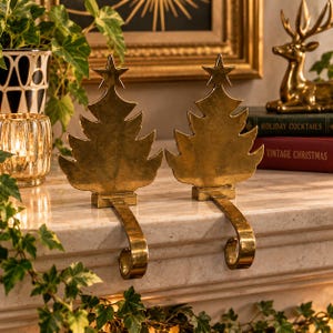 May include: A pair of gold-toned metal stocking holders, each shaped like a stylized Christmas tree topped with a star. The flat tree silhouettes are mounted on rectangular bases with curved, hook-shaped arms designed to hang over a ledge.