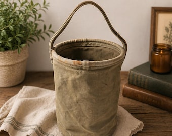 May include: An olive green canvas bucket with a matching handle. The bucket sits on a cream-colored cloth with gray stripes. A potted plant, books, and a candle in a jar are in the background.