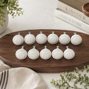 May include: A collection of twelve white ceramic cabinet knobs with silver-toned stems, arranged on a dark brown wooden oval serving board. The knobs have a simple, rounded design. The background includes books and greenery.