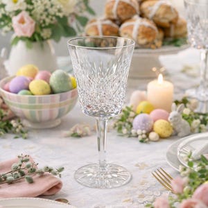 May include: A clear crystal wine glass with a detailed cut pattern stands on a table set for Easter. The table is decorated with pastel-colored Easter eggs, a bowl of eggs, flowers, and hot cross buns. A lit candle and a small bunny figurine are also present.