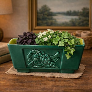 Puede incluir: Una jardinera rectangular de cerámica verde oscuro con un diseño en relieve de un pájaro y uvas. La jardinera está llena de una variedad de plantas, incluyendo follaje morado oscuro, blanco y verde. La jardinera se encuentra sobre una estera de arpillera.