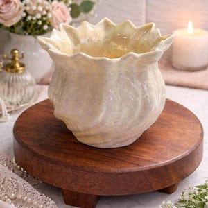 May include: A cream-colored, seashell-shaped ceramic bowl sits on a round, dark wooden tray. The bowl has a textured surface and a ruffled rim. The tray is elevated by small feet. A lit candle and flowers are in the background.