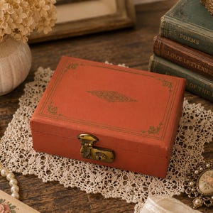 May include: A rectangular, terracotta-colored storage box featuring a gold-toned metal latch closure on the front. The lid is decorated with a thin, gold-colored border and a central, intricate, elongated floral-inspired filigree design.