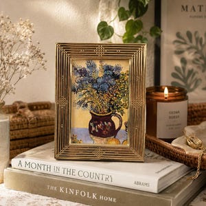 May include: A rectangular gold-toned metal picture frame with a textured, linear border pattern. The frame contains a print of a floral still life painting featuring blue flowers in a dark pitcher on a yellow background, resting on a stack of books.