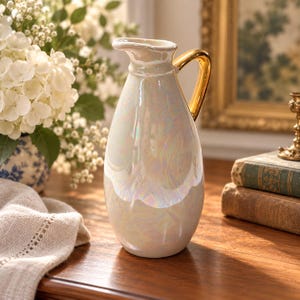 May include: A pearlescent white ceramic pitcher featuring a smooth, iridescent finish that reflects soft colors. The pitcher has a curved spout and a polished gold-tone handle attached to the side, creating a contrast with the glossy body.