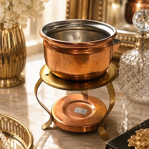 May include: A copper pot with a silver-toned interior sits atop a circular brass stand with three curved legs. The base of the stand features a small rectangular label with text. The copper surface shows signs of wear and natural patina.