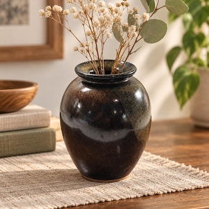 May include: A rounded ceramic vase with a dark, mottled black and brown glaze featuring a glossy finish. The vase has a wide, short neck and a flared rim, containing a small arrangement of dried white flowers and green eucalyptus leaves.