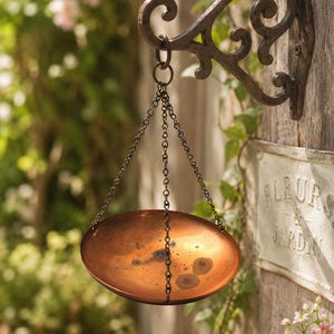 May include: A hanging copper bowl suspended by three thin metal chains from a dark, ornate, scroll-patterned iron wall bracket. The bowl has a warm, metallic finish with subtle dark spots and a smooth, rounded shape, designed for wall mounting.