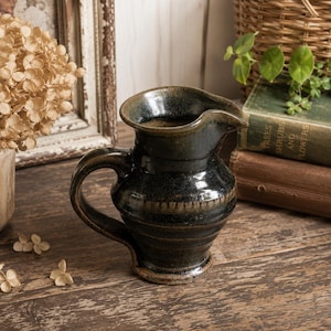May include: A dark, mottled green and brown ceramic pitcher with a rounded body, a wide pouring spout, and a curved handle. The surface features horizontal ridges and a glossy glaze finish, showcasing a deep, earthy color palette throughout.