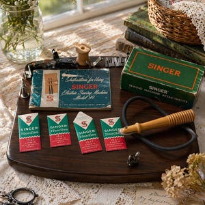 May include: A vintage sewing kit with Singer needles and attachments. The kit includes a green box, needle packets, and a manual for a Singer electric sewing machine. The items are arranged on a dark wooden tray.