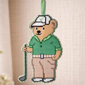 May include: A cross-stitch bear ornament dressed in a green golf shirt, khaki trousers, and a white cap. The bear holds a golf club and has a green ribbon for hanging. The ornament features black stitching and a white belt.