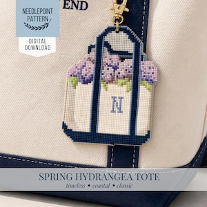 Op de afbeelding: Een close-up van een witte tote bag met marineblauwe rand en een goudkleurige sleutelhanger. De sleutelhanger is een borduurontwerp van een tote bag gevuld met paarse hortensia's en de letter "N". De tekst "SPRING HYDRANGEA TOTE" staat onderaan.