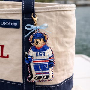 Könnte beinhalten: Ein handgefertigter Schlüsselanhänger mit einem Bären in einem USA-Hockey-Trikot in Blau, Weiß und Rot. Der Bär hält einen Hockeyschläger. Der Schlüsselanhänger ist an einer Lands' End-Tragetasche mit blauem Riemen und goldenem Verschluss befestigt.