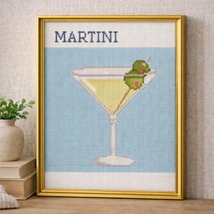 Op de afbeelding: Een ingelijst kruissteekkunstwerk met een martini-cocktail. Het woord "MARTINI" is bovenaan gestikt. Het cocktailglas is gevuld met een gele vloeistof en gegarneerd met twee groene olijven op een cocktailprikker. De achtergrond is lichtblauw.