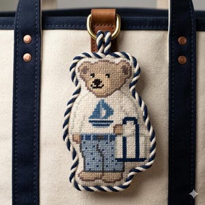 Puede incluir: Un llavero de oso de peluche bordado a punto de cruz con temática náutica. El oso viste una camisa blanca con un velero azul y pantalones azules estampados. El llavero está ribeteado con una cuerda azul y blanca y sujeto a una correa de cuero.