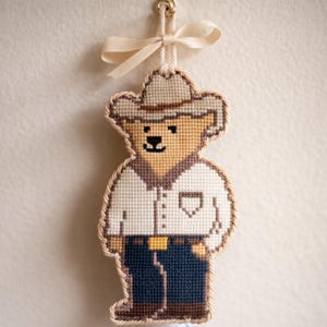 May include: A handmade, cross-stitched ornament in the shape of a bear wearing a cowboy hat and outfit. The bear has a tan face and hat, a white shirt, blue jeans, and a brown belt. A beige ribbon is attached for hanging.