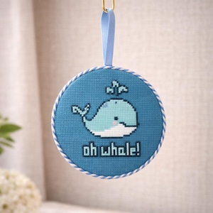 May include: A round, blue cross-stitch ornament featuring a light blue whale with a water spout. The text "oh whale!" is below the whale. The ornament has a blue ribbon for hanging and a white and blue rope border.