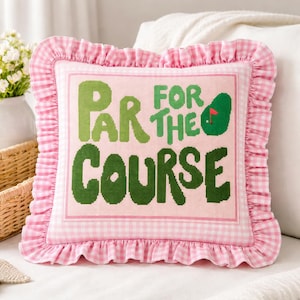 Könnte beinhalten: Ein quadratisches Zierkissen mit einem rosa Vichy-Karo-Rand und Rüschenbesatz. Das Kissen zeigt den Text "PAR FOR THE COURSE" in grünen Buchstaben auf rosa Hintergrund, mit einer Golfball-Grafik.